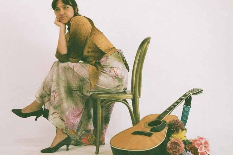 Arielle Soucy poses in a chair. She wears a floral skirt, and a guitar rests over a bouquet of flowers on the floor beside her.